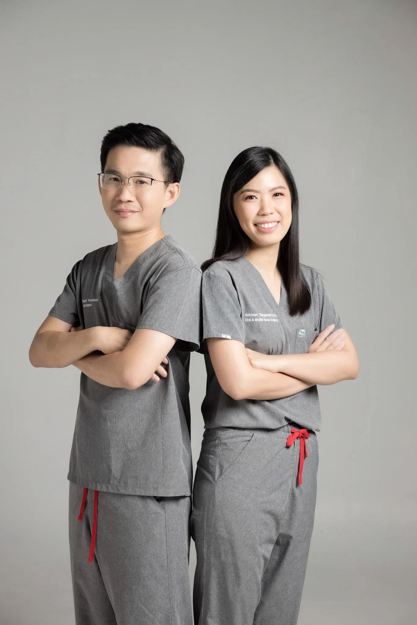 ทันตแพทย์ชายและหญิงของ The Area Plus Dental Clinic ในชุดสครับสีเทา ยืนไขว้แขนหันหลังชนกัน
